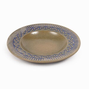 Puede incluir: Un plato redondo de cerámica con un centro poco profundo y un borde elevado. El plato es de color beige con un patrón geométrico azul en el borde. El plato probablemente se utiliza para servir alimentos.
