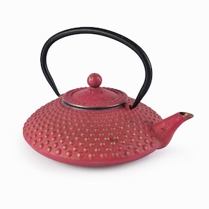 Könnte beinhalten: Eine rote gusseiserne Teekanne mit einem schwarzen Griff und einer strukturierten Oberfläche. Die Teekanne hat einen runden Deckel mit einem kleinen Knopf oben.
