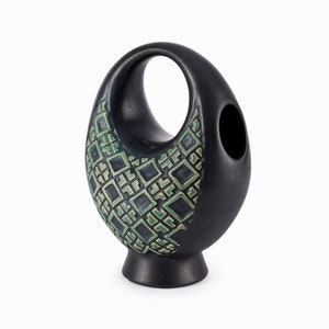 Puede incluir: Jarrón de cerámica negro con un diseño geométrico verde y negro. El jarrón tiene una forma única con una gran abertura en la parte superior y una abertura más pequeña en el lateral.