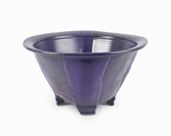 May include: A purple ceramic planter with a wide, rounded rim and three short legs. The planter has a textured, slightly speckled surface.