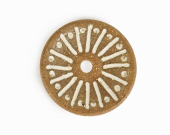 May include: A brown ceramic disc with a white sunburst design. The disc has a hole in the centre.