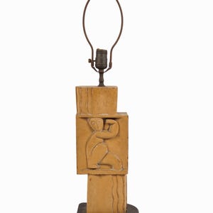 May include: A tan-colored table lamp with a stacked rectangular design. The lamp features a carved relief of a person. The lamp has a brown metal lamp shade holder and sits on a black base.