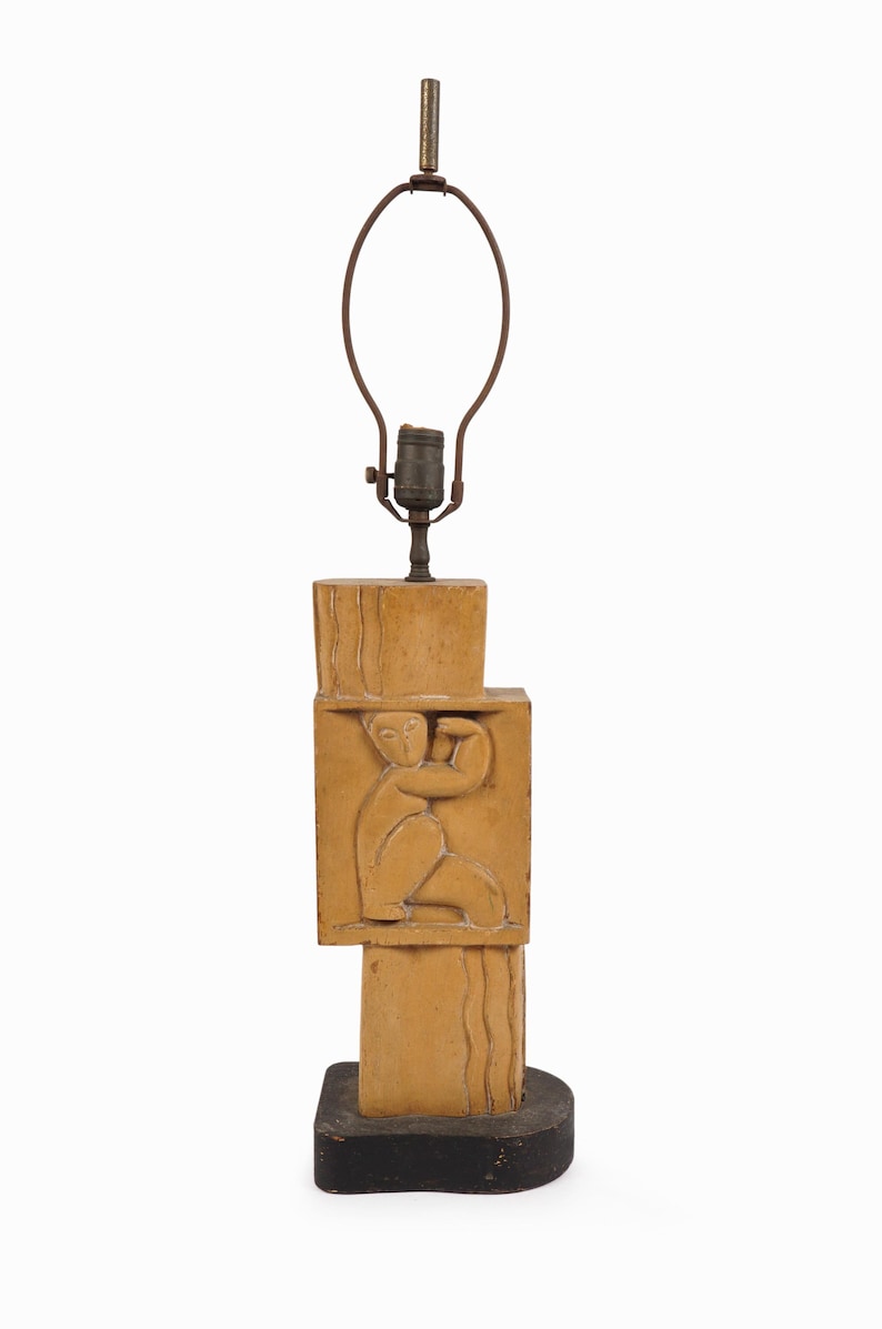 May include: A tan-colored table lamp with a stacked rectangular design. The lamp features a carved relief of a person. The lamp has a brown metal lamp shade holder and sits on a black base.