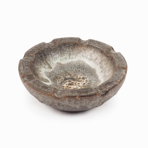 May include: A handcrafted, ceramic ashtray with a unique, organic shape. The ashtray features a mottled, earthy color palette of gray, brown, and white, with a textured surface. The design includes several notches for resting cigarettes.