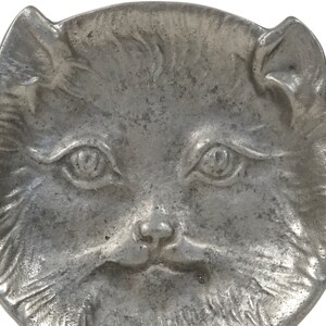 Puede incluir: Un plato de metal plateado en forma de cara de gato. El plato tiene un dise&ntilde;o en relieve detallado de una cara de gato con ojos grandes, una nariz peque&ntilde;a y una boca ligeramente abierta.