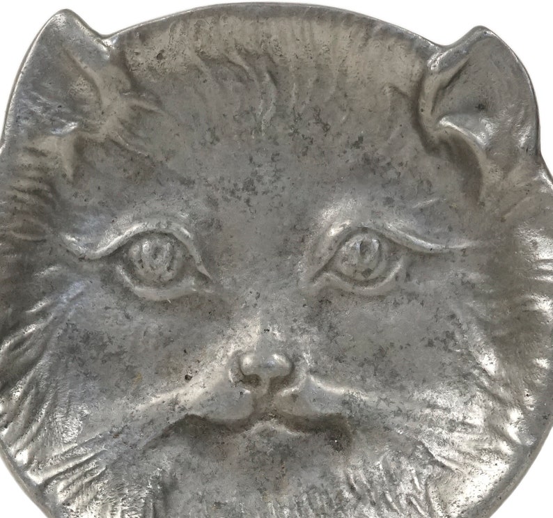 Puede incluir: Un plato de metal plateado en forma de cara de gato. El plato tiene un dise&ntilde;o en relieve detallado de una cara de gato con ojos grandes, una nariz peque&ntilde;a y una boca ligeramente abierta.