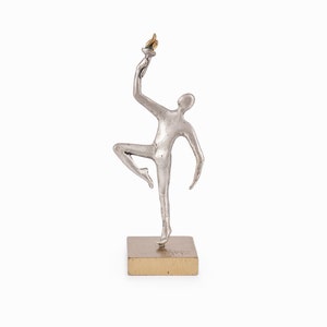 May include: A silver sculpture of a person holding a torch, standing on a gold base. The sculpture is abstract and stylized.
