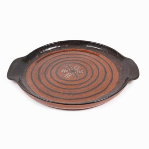 Puede incluir: Una bandeja de servir de cerámica marrón con dos asas. La bandeja tiene un borde negro y un diseño de círculos concéntricos con un patrón floral estilizado en el centro.