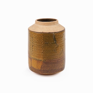 May include: A cylindrical ceramic jar with a wide opening and a slightly tapered neck. The jar features a textured, earthy design with shades of brown and tan. The lower portion is a darker brown, and the upper section is a lighter tan color.