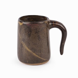May include: A brown ceramic mug with a unique, curved handle. The mug has a speckled, textured finish.