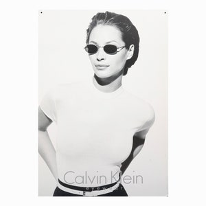 May include: Black and white photo of a woman wearing oval-shaped sunglasses and a white t-shirt. The text "Calvin Klein eyewear" is printed below the woman.