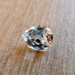 May include: A clear pear-shaped gemstone with facets, on a wood background.