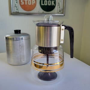 Peut inclure: Une cafetière à percolation vintage en verre et métal avec des accents dorés et une poignée foncée. Une boîte argentée avec un couvercle noir est à gauche. Un panneau encadré au-dessus indique "STOP LOOK".
