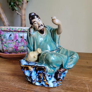 Peut inclure: Figurine en céramique d'un homme assis en robes turquoise, tenant une petite coupe. La figurine a une longue barbe et est assise sur une base bleue et marron. Une petite cruche beige repose à côté de lui. La figurine est probablement d'origine asiatique.