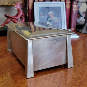 May include: A silver rectangular box with a raised lid and a smooth, polished surface. The box has four legs and a slightly angled design.