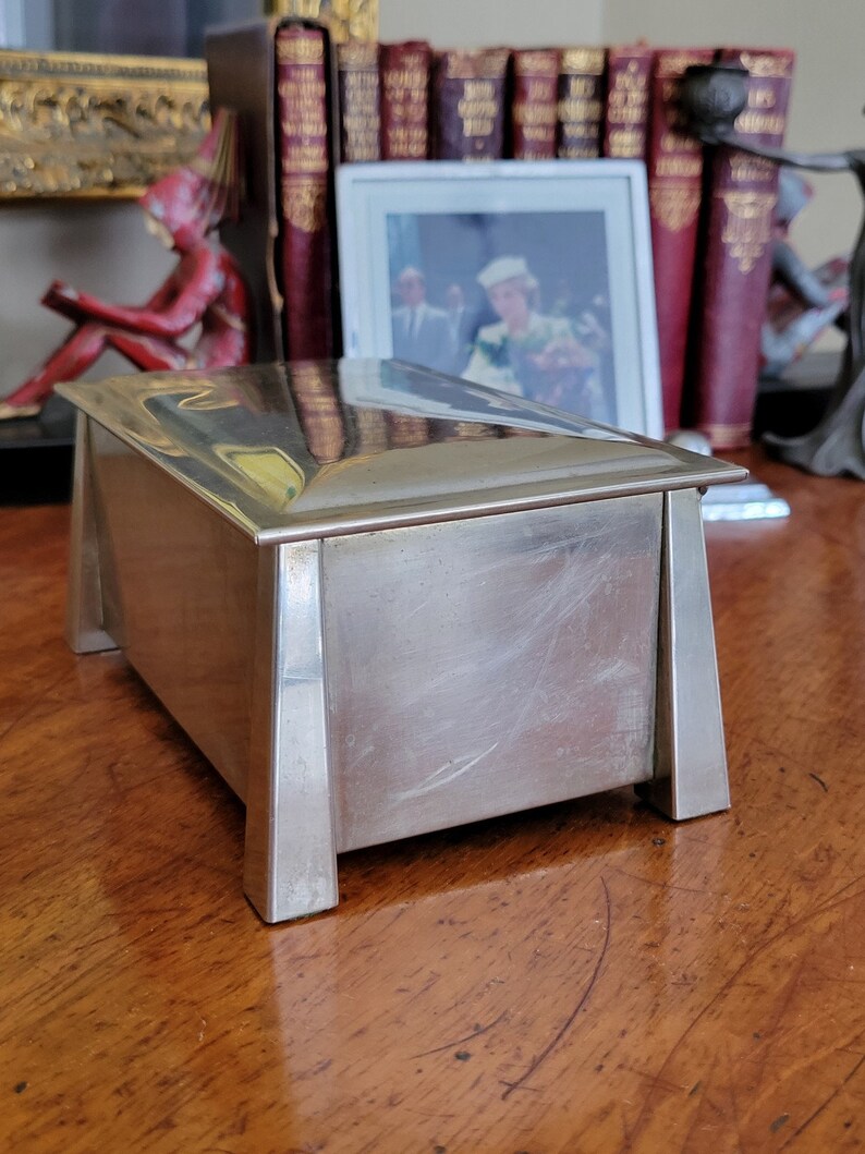 May include: A silver rectangular box with a raised lid and a smooth, polished surface. The box has four legs and a slightly angled design.