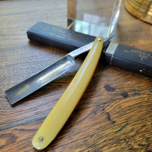 May include: A classic straight razor with a pale yellow handle and a silver blade, displayed open on a wooden surface. The razor's sharp edge is visible. Two black cases are in the background.