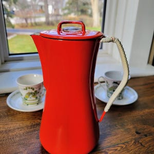 May include: A bright red enamel coffee pot with a lid and a white woven handle. The pot is on a wooden surface, with two teacups and saucers in the background. The coffee pot is approximately 25 cm tall.
