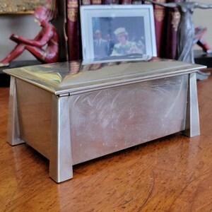 May include: A silver rectangular box with a hinged lid and a polished finish. The box has a simple, geometric design with squared corners and a raised edge around the lid. The box is sitting on a wooden surface.