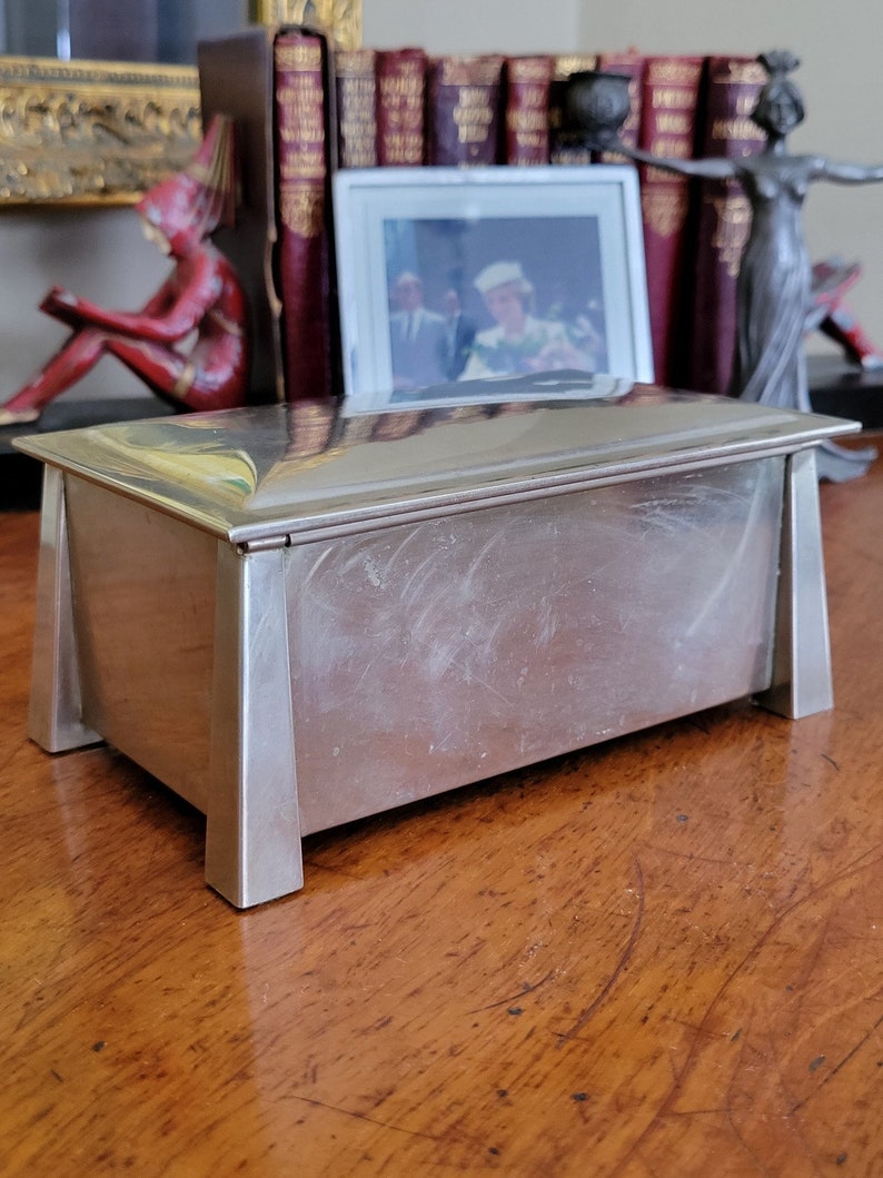 May include: A silver rectangular box with a hinged lid and a polished finish. The box has a simple, geometric design with squared corners and a raised edge around the lid. The box is sitting on a wooden surface.