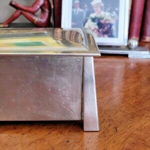 May include: A silver rectangular box with a slightly raised lid. The box has a smooth, polished surface with some scratches. The box is sitting on a wooden surface.