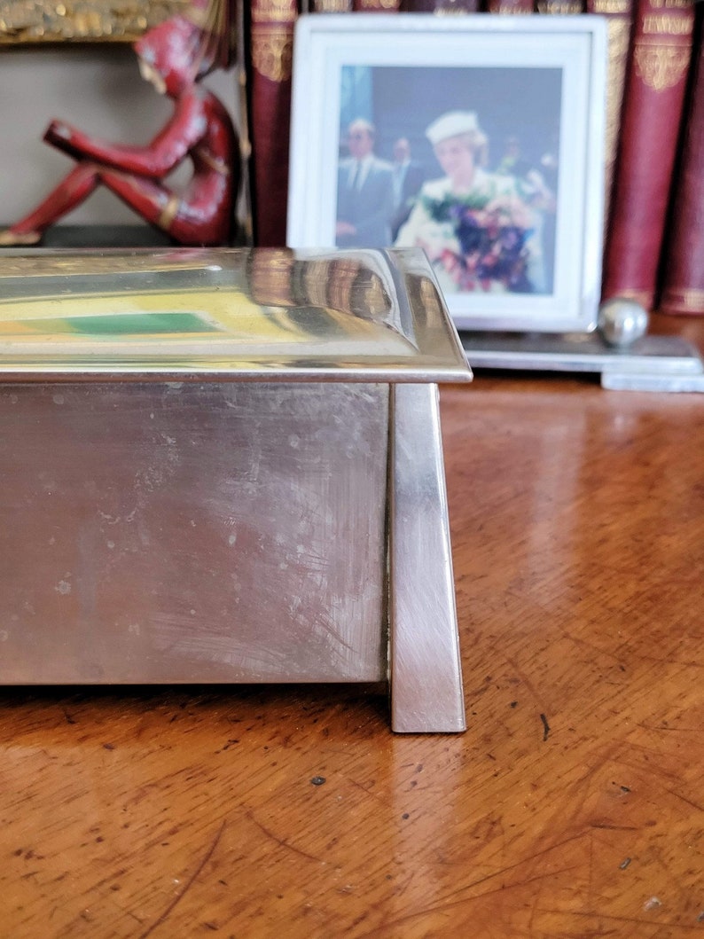 May include: A silver rectangular box with a slightly raised lid. The box has a smooth, polished surface with some scratches. The box is sitting on a wooden surface.