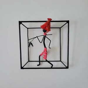 May include: A black wire sculpture of a person wearing a red and white polka dot skirt and a red hat. The sculpture is inside a black wire cube.