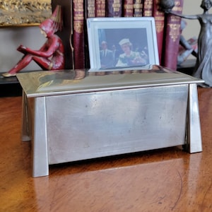 May include: A silver rectangular box with a raised lid and a slightly textured surface. The box is sitting on a wooden surface.