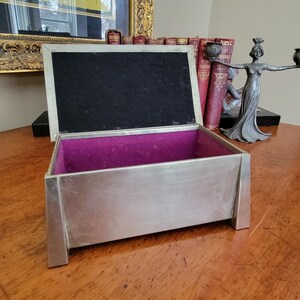 May include: A silver metal box with a purple velvet interior. The box is open and the lid is propped up. The box is sitting on a wooden surface.