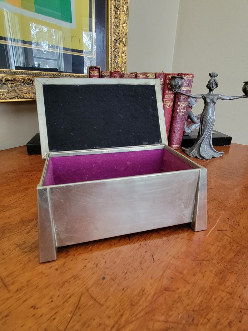 May include: A silver metal box with a purple velvet interior. The box is open and the lid is propped up. The box is sitting on a wooden surface.