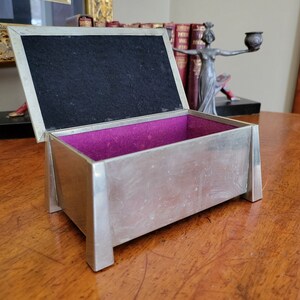 May include: A silver rectangular box with a purple velvet interior and a black felt lid. The box is open and sitting on a wooden surface.