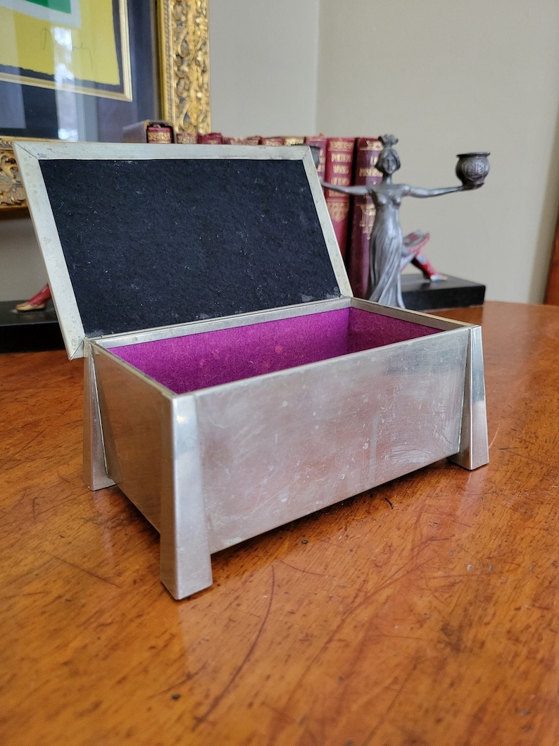 May include: A silver rectangular box with a purple velvet interior and a black felt lid. The box is open and sitting on a wooden surface.
