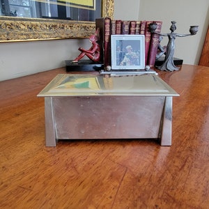 May include: A silver metal box with a hinged lid, sitting on a wooden surface. The box has a rectangular shape and a smooth, reflective surface.