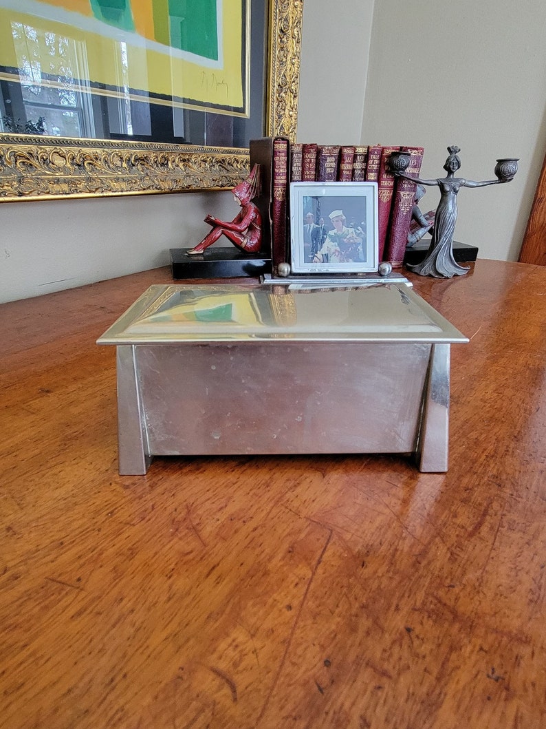 May include: A silver metal box with a hinged lid, sitting on a wooden surface. The box has a rectangular shape and a smooth, reflective surface.