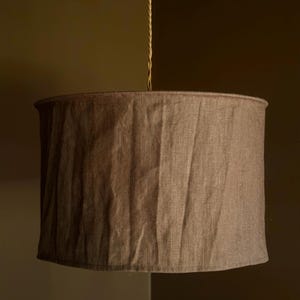 May include: A brown fabric lampshade suspended from a beige cord. The lampshade has a cylindrical shape with a slightly curved bottom edge. The fabric appears to be textured, with visible vertical folds. The background is a muted brown.