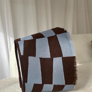 May include: A folded textile featuring a brown and light blue chequered pattern. The fabric, possibly a throw or decorative item, displays a repeating design of rectangular blocks. The edges have a fringed finish.