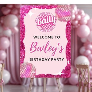 Op de afbeelding: Een roze glitter welkomstbord voor Bailey's verjaardagsfeest. Het bord heeft de tekst "Welcome to Bailey's Birthday Party" in een cursief lettertype, met een discobal afbeelding. Het bord staat op een houten ezel, met roze ballonnen op de achtergrond.