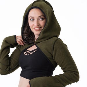 May include: An olive green hooded shrug with black trim, paired with a black crop top and black leggings with a sheer panel. The shrug features long sleeves and a hood. The outfit is designed for activewear.