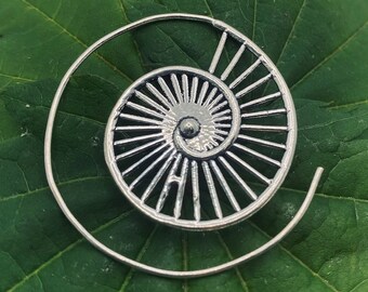 May include: A silver-colored spiral earring with a detailed, openwork design resembling a nautilus shell. The earring is set against a green leaf, highlighting its intricate pattern and metallic sheen. The spiral measures approximately 1.5 inches in diameter.