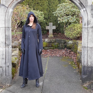 May include: A woman wearing a long, black hooded coat stands in front of a stone archway. The archway is decorated with a carved stone cross. The woman is wearing black boots and has her hair pulled back.