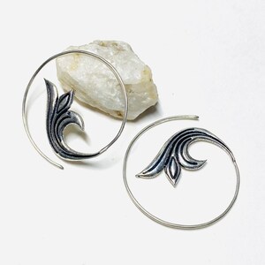 Peut inclure: Une paire de boucles d'oreilles spirales argentées. Chaque boucle d'oreille présente un motif de feuille stylisé de couleur foncée. Les boucles d'oreilles sont présentées sur une surface blanche avec une roche de couleur claire en arrière-plan.