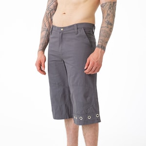 May include: Grey capri trousers with metal eyelet detailing at the hem. The capris have a button closure and pockets. The model is wearing black leather ankle boots. The trousers are made of a durable fabric.