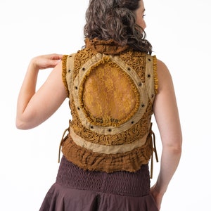 Op de afbeelding: Een bruin en beige vest zonder mouwen met een rugpaneel van kant en een bruine rok. Het vest heeft een kantontwerp en wordt in de taille gebonden.