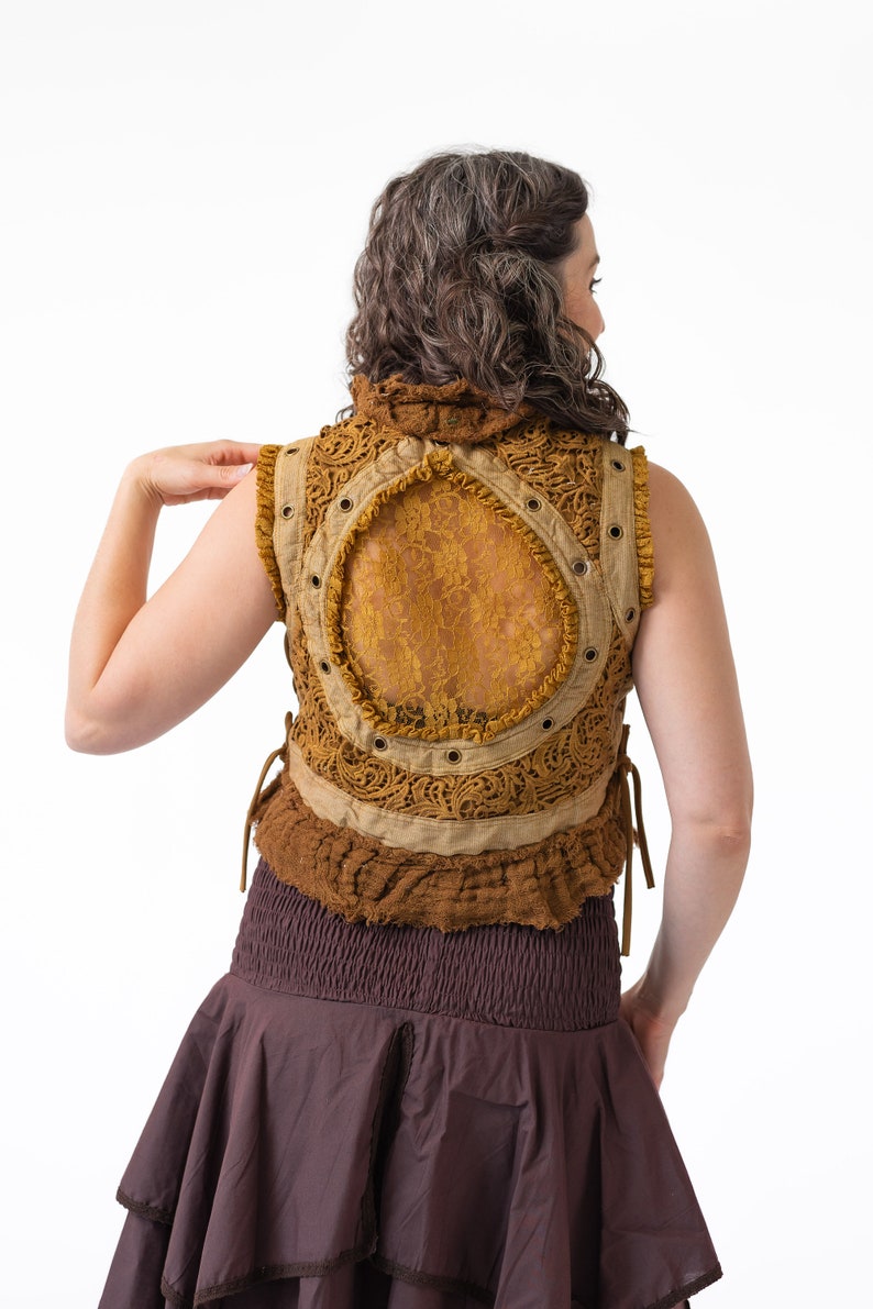 Op de afbeelding: Een bruin en beige vest zonder mouwen met een rugpaneel van kant en een bruine rok. Het vest heeft een kantontwerp en wordt in de taille gebonden.