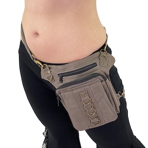 May include: A brown canvas fanny pack with a zippered pocket and multiple metal rings. The fanny pack is worn on a person's hip.