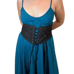 May include: A black corset belt with a lace-up front and eyelets. The belt is worn over a teal dress. The corset belt is a wide, structured piece with a defined shape, and it appears to be made of a sturdy fabric.