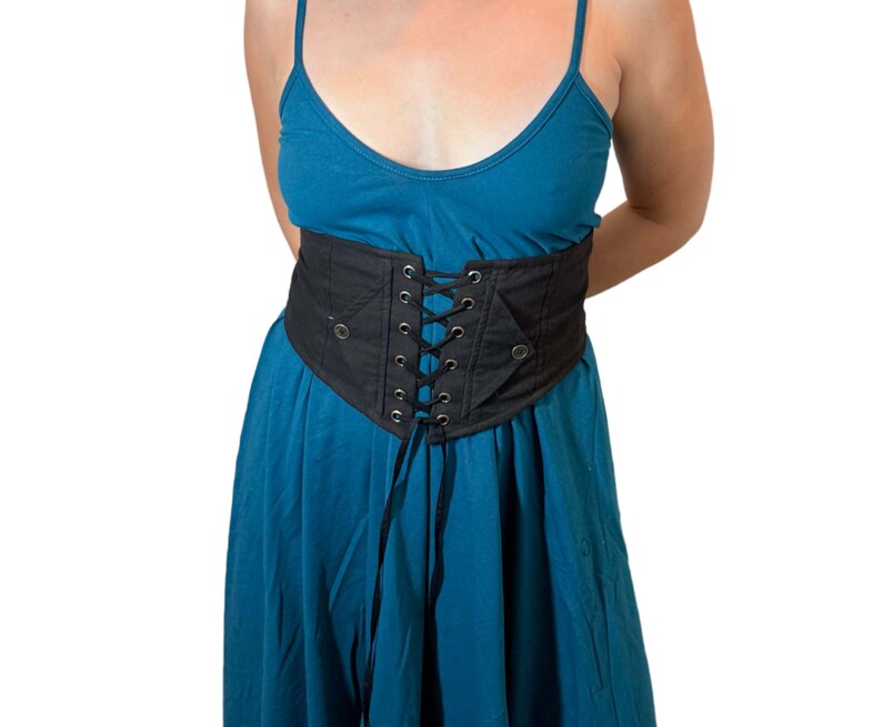 May include: A black corset belt with a lace-up front and eyelets. The belt is worn over a teal dress. The corset belt is a wide, structured piece with a defined shape, and it appears to be made of a sturdy fabric.