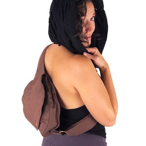 May include: A brown canvas crossbody bag with a quilted design. The bag has a long strap that goes over the shoulder and a smaller pocket on the front. The bag is worn by a person with a black hooded top.