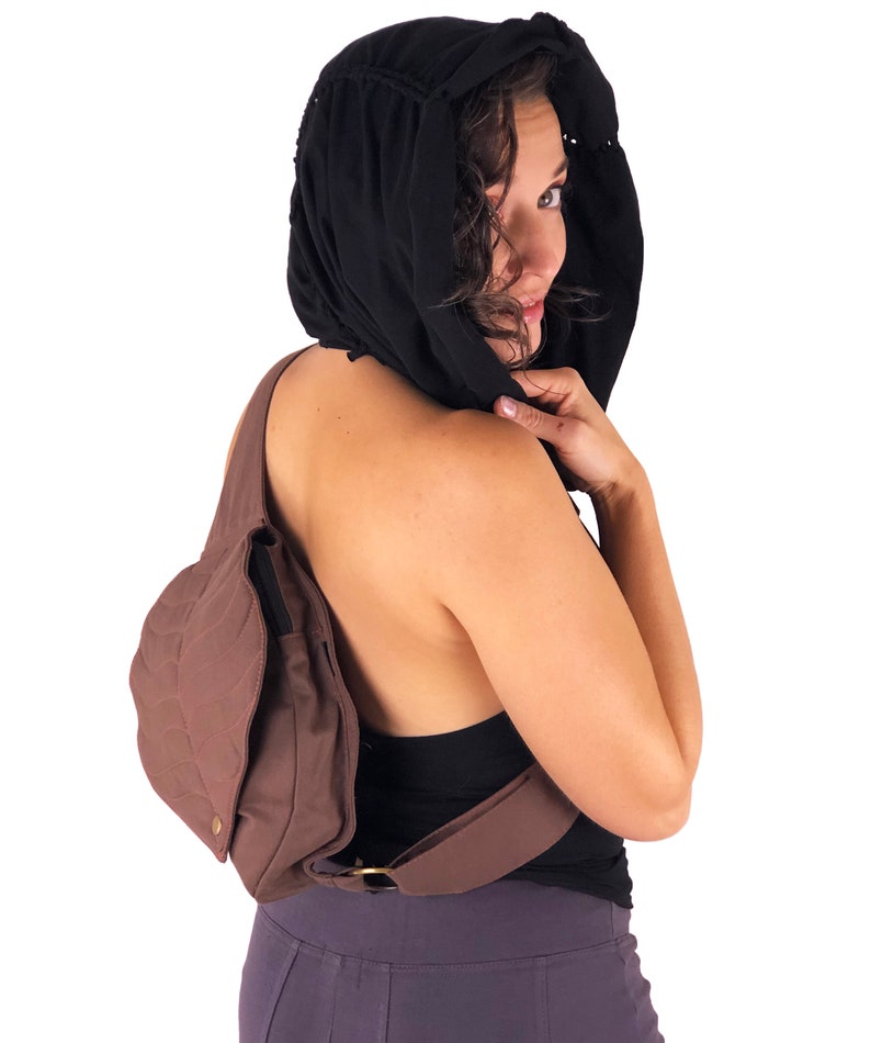 May include: A brown canvas crossbody bag with a quilted design. The bag has a long strap that goes over the shoulder and a smaller pocket on the front. The bag is worn by a person with a black hooded top.
