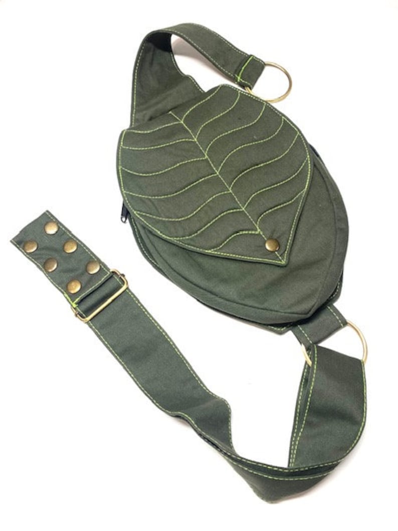 May include: A green canvas crossbody bag with a leaf design. The bag has a long adjustable strap and a metal buckle. The leaf design is stitched with a yellow thread.